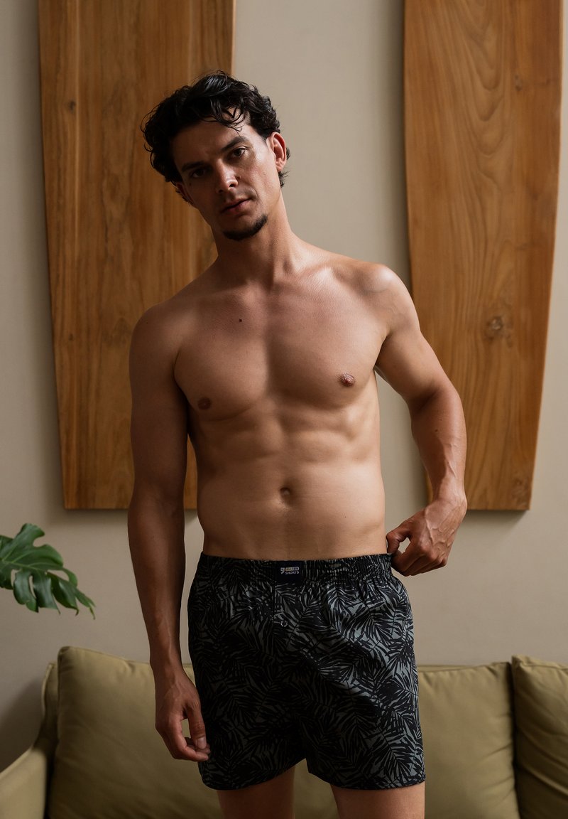 Zwarte boxers met een bladmotief, gemaakt van zacht stof, met een elastische tailleband. Model met een gespierd postuur, staand binnen.