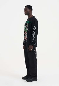 Schwarzes Langarmshirt mit grafischem Aufdruck und grüner Schrift, kombiniert mit schwarzen Hosen mit einem Webmuster. Getragen mit schwarzen Turnschuhen.
