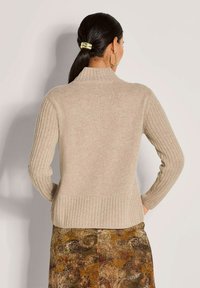 Frau mit dunklen Haaren im Pferdezopf, die große goldene Creolen, einen beigen gerippten Pullover und einen gemusterten braunen Rock trägt und von hinten zu sehen ist.