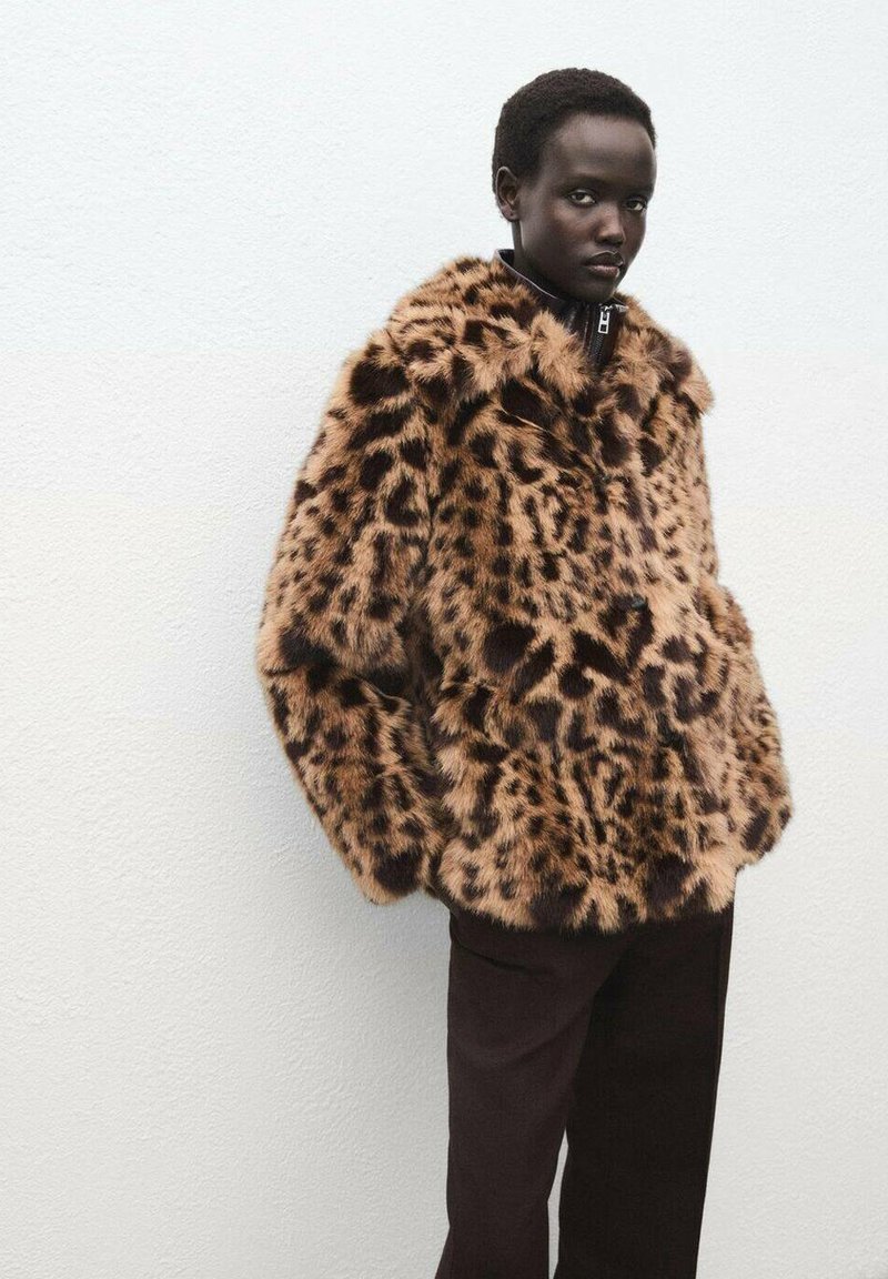 Giacca in pelliccia sintetica con stampa leopardata marrone, dal tessuto spesso e morbido, con zip sul davanti, indossata su pantaloni scuri. Sfondo semplice.