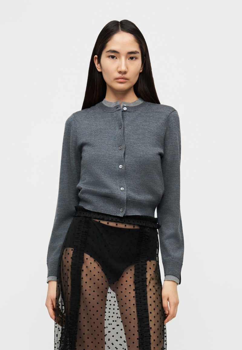 Γκρι ζακέτα με στρογγυλή λαιμόκοψη, μακριά μανίκ ια και κλείσιμο με κουμπιά, συνδυασμένη με διάφανο μαύρο φούστα με πουά και διακοσμητικές βολάν.