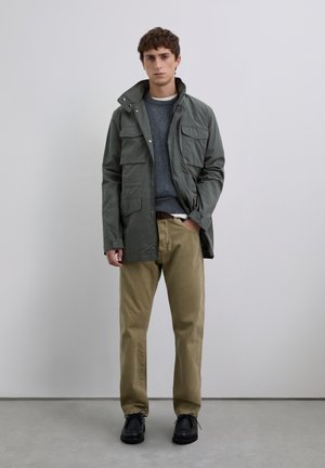 Junger Mann in einer grünen Feldjacke, grauem Pullover, beigen Hosen und schwarzen Schuhen, der vor einem schlichten hellgrauen Hintergrund steht.