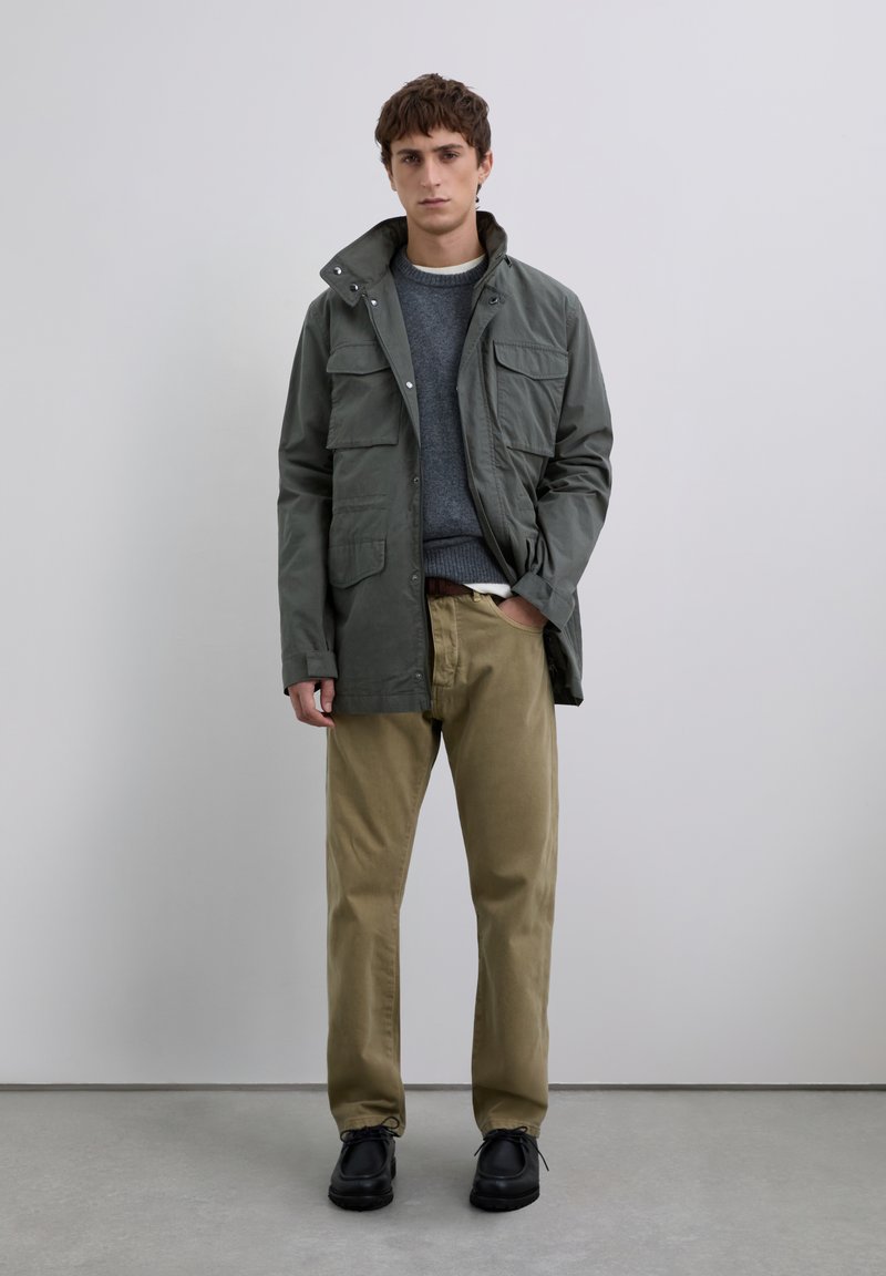 Young man wearing green field jacket, gray sweater, beige pants, and black shoes standing against plain light gray background.