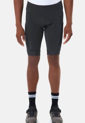 Uomo che indossa pantaloncini da ciclismo neri, calzini neri con strisce bianche e scarpe da ciclismo grigie, in piedi davanti a uno sfondo bianco.