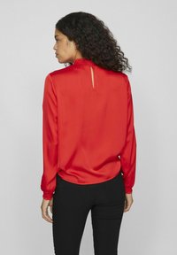 Blouse en satin rouge avec un col haut orné de volants, des manches longues avec des poignets élastiques et une ouverture en forme de clé au dos.
