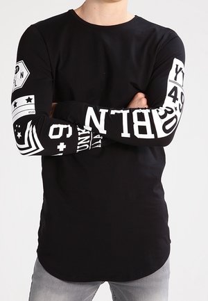 Personne portant un t-shirt noir à manches longues avec des chiffres et lettres blancs sur les manches, bras croisés, pantalon gris clair.