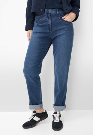 Person iført opsmøgede blå jeans, sorte og hvide sneakers samt en mørk jakke med den ene hånd i lommen, stående foran en hvid baggrund.