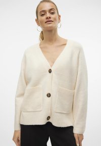 Lys cremefarvet cardigan med V-hals, ribbede manchetter, to frontlommer og dekorative knapper, lavet af et blødt, tekstureret stof.