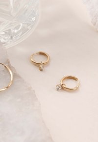 Goldene Creolenohrringe mit einem kleinen klaren Steinakzent. Glatte, glänzende Oberfläche mit runder, hohler Form, vor einem hellen Hintergrund.