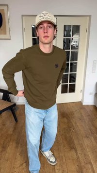 Νεαρός άνδρας που φοράει μπεζ καπέλο, χακί φούτερ, ανοιχτό μπλε τζιν και αθλητικά παπούτσια στέκεται σε ένα δωμάτιο με ξύλινο πάτωμα και πόρτα με γυάλινα πάνελ.