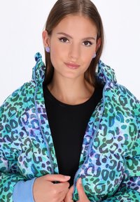Giacca imbottita in tessuto leopardato verde e blu, con chiusura a zip, cappuccio e polsini a costine blu chiaro. Camicia nera sotto.