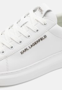 Fehér bőrsneaker kerek formával, fehér fűzőkkel, ezüst fémlyukakkal és fényes logóval az oldalon. Simogatós textúra.