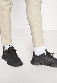Zapatillas deportivas negras con parte superior de malla, tres rayas horizontales, suelas acolchadas y cordones negros, combinadas con pantalones de chándal ajustados de color crema.