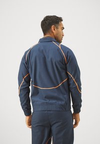Mornarsko modra atletska jakna z oranžnim kontrastnim šivom, ovratnikom in elastičnimi manšetami. Lahkoten material z gladko teksturo.