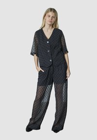 Ung kvinde står med hænderne i lommerne, iført en gennemsigtig sort top med prikker og bukser over en sort underlayer mod en ensfarvet baggrund.