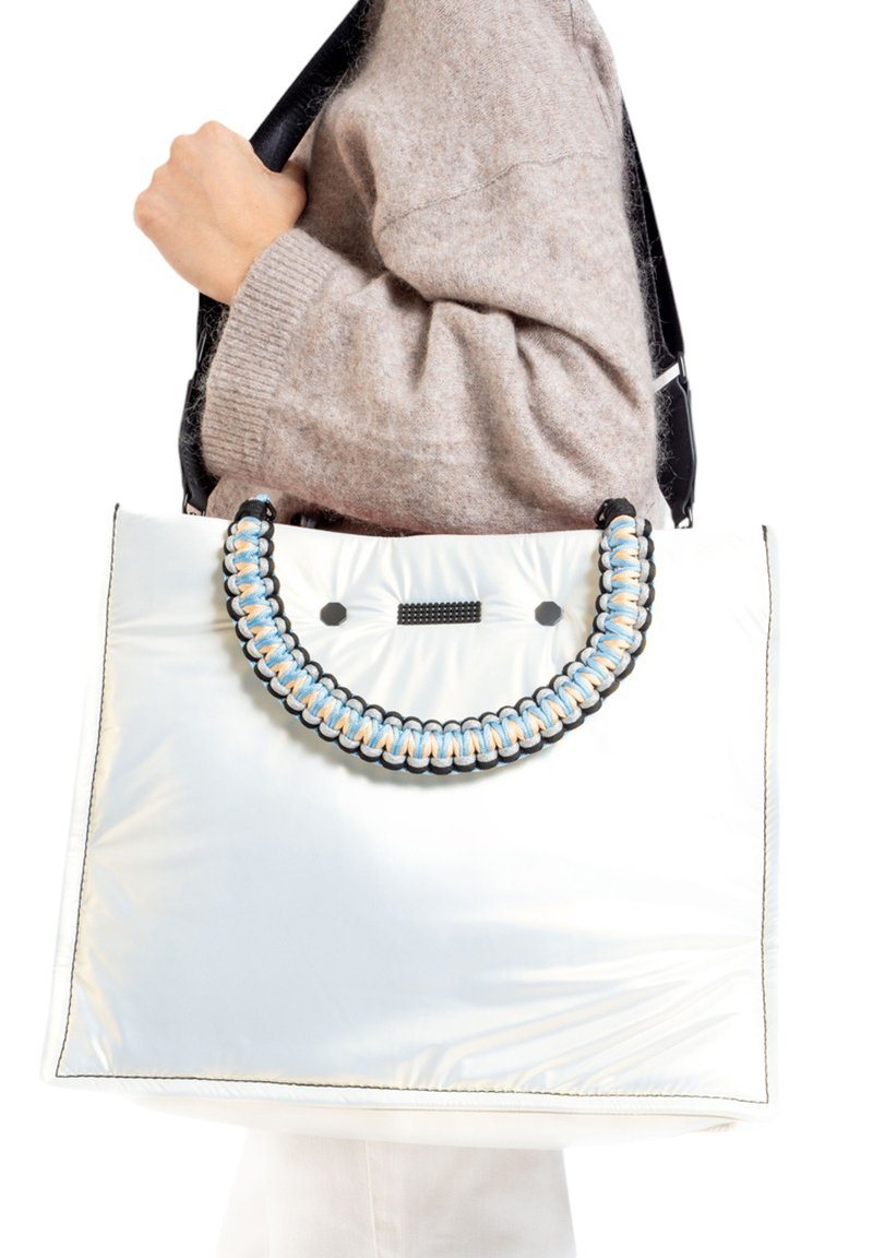 Grand sac fourre-tout blanc en matériau brillant, avec une poignée tressée ornée d'accents noirs, bleus et orange. Texture lisse, détails minimaux.