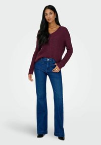Maglione lavorato a maglia bordeaux con scollatura a V e maniche lunghe, abbinato a jeans a campana blu scuro con tasche frontali e chiusura a bottone.