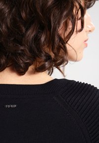 Černý žebrovaný svetr s kulatým výstřihem. Texturované akcenty na švech ramen a malé logo "RAW" na zadním výstřihu.