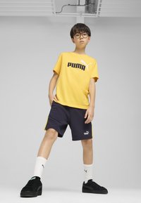 Gul bomullströja med "PUMA"-logotyp, tillsammans med marinblå shorts med gula detaljer, vita strumpor och svarta sneakers.