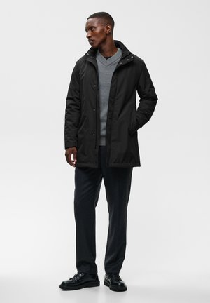 Homme portant un manteau noir par-dessus un pull gris à col en V, un pantalon foncé et des chaussures noires, debout avec une main dans la poche devant un fond uni.