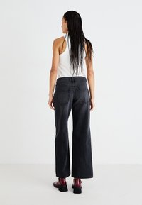 Jeans de mezclilla negra de pierna ancha con una cinturilla de altura media, dos bolsillos traseros y una textura suave, combinados con zapatos burdeos brillantes.