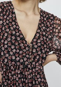 Robe fleurie avec un fond noir, ornée de fleurs roses, décolleté en V profond et boutons sur le devant. Texture plissée sur toute la surface.