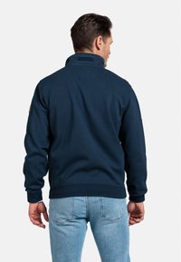 Marineblauwe fleecetruien met een hoge kraag, geribbelde manchetten en onderkant, en een patch op de linkerarm, gecombineerd met lichtblauwe jeans.