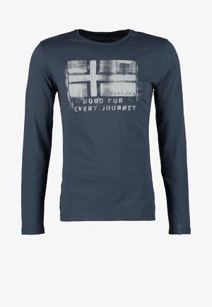 Mann trägt schwarze Lederjacke, dunkle Jeans, weinrote Sneakers, blaues Grafik-T-Shirt, hält eine graue Tragetasche mit schwarzen Trägern.