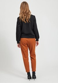 Vrouw met golvend blond haar in een zwarte lange mouwtop, roestkleurige tapered broek en zwarte hoge enkellaarsjes, met de rug naar de camera gekeerd.