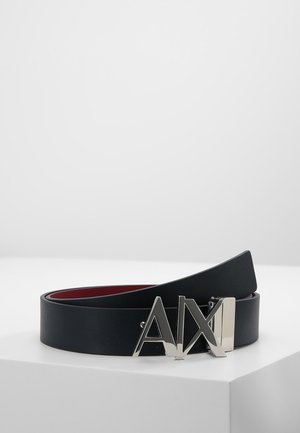 Cintura in pelle nera con fibbia in metallo argento "AX", arrotolata e posata su una superficie bianca con uno sfondo grigio chiaro uniforme.