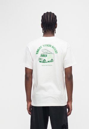 Homme aux cheveux tressés portant un t-shirt blanc avec un motif vert d'un camion de glaces et le texte "Treat Your Sole!" dans le dos.