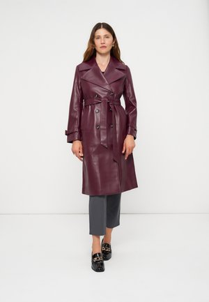 Femme debout dans un trench en faux cuir bordeaux avec ceinture, pantalon gris et mocassins noirs sur fond blanc uni.