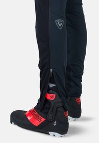 Schwarze Skihosen mit Reißverschlüssen an den Knöcheln, kombiniert mit schwarz-roten Skischuhen, die ein auffälliges Logo und verstellbare Riemen besitzen.