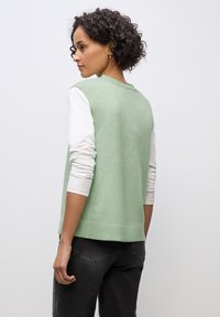 Zielony bezrękawnik z teksturowanym splotem, nałożony na białą koszulę z długimi rękawami, w połączeniu z ciemnymi dżinsami.