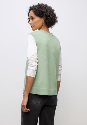Groene mouwloze trui met een textuur brei-ontwerp, gelaagd over een witte langemouwen shirt, gecombineerd met een donkere spijkerbroek.
