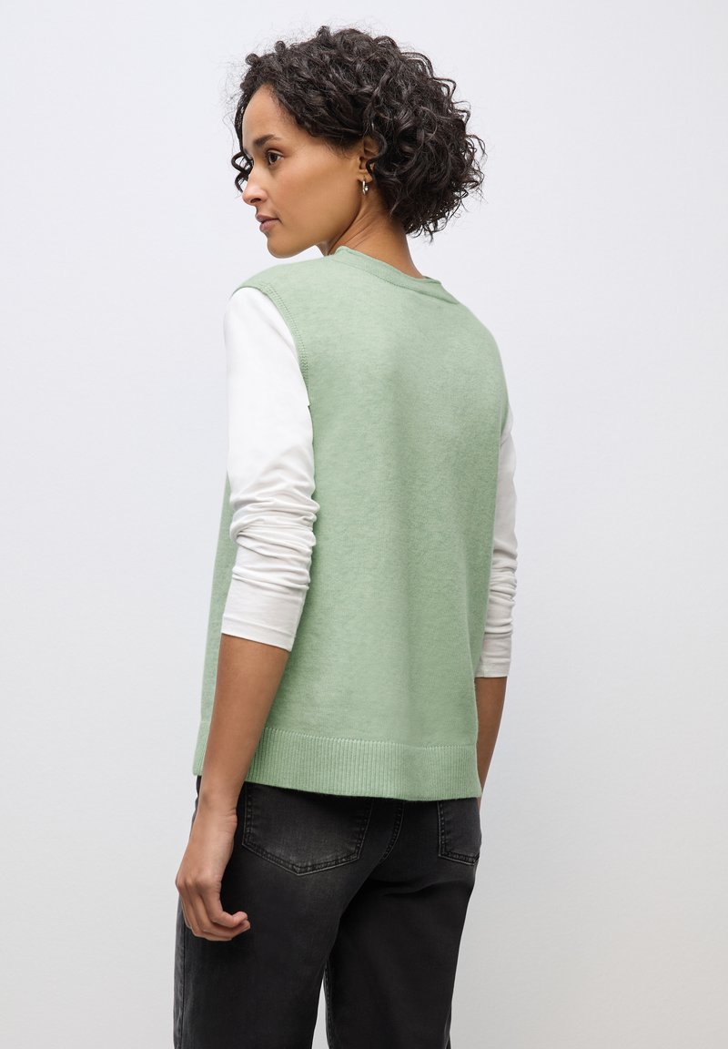 Zielony bezrękawnik z teksturowanym splotem, nałożony na białą koszulę z długimi rękawami, w połączeniu z ciemnymi dżinsami.