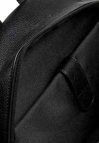 Innenansicht eines schwarzen Leder-Rucksacks mit glattem Stofffutter. Er verfügt über einen Reißverschluss und eine Seiteninnentasche mit Druckknopfverschluss.