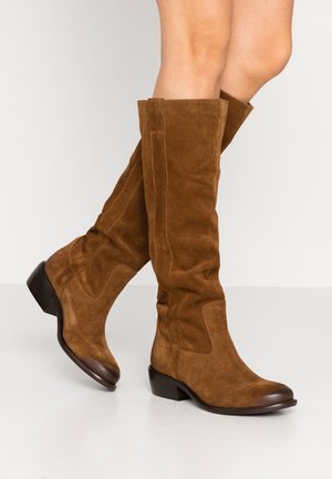 Jambes portant des bottes hautes en suède marron avec des talons blocs bas, debout sur un sol clair sur fond uni.