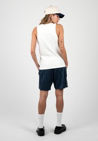 Femme vêtue d'un débardeur blanc, d'un short rayé bleu marine, de chaussettes blanches, de chaussures noires et d'une casquette de baseball beige, debout avec les mains dans les poches, de dos.