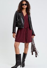 Czarna skórzana kurtka typu cropped na bordowej sukience z delikatnym zwierzęcym wzorem. Czarne kozaki do kolan na obcasie i mała torebka z frędzlami.