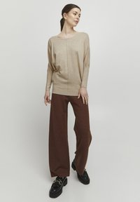 Beige Oversize-Pullover mit U-Boot-Ausschnitt und gestrickter Textur, kombiniert mit weiten braunen Hosen und schwarzen Plateauschuhen mit goldenen Beschlägen.