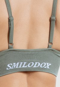 Olijfgroene bralette met ribbelstructuur en verstelbare bandjes. Voorzien van het witte "SMILODOX" logo over de band. Achteraanzicht.