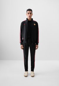 Black tracksuit with red stripes; zip-up jacket featuring logo and patches, paired with pants. Accent sneakers in cream and red.