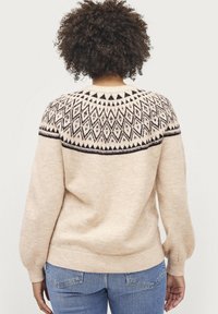 Pull en tricot beige avec un motif géométrique noir et blanc sur l'empiècement, doté de manches ballon et d'un ourlet côtelé. Texture douce.