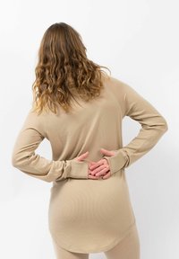 Beige langwerpige top met een ribbelstructuur, voorzien van een high-low zoom en duimgaten, gedragen door een persoon met golvend haar en handen op de heupen.