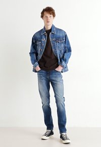 Jachetă din denim de culoare albastră deschisă, tricou maro, blugi albaștri decolorati și teniși negri cu glezna înaltă. Modelul stă cu mâinile în buzunare pe un fundal neutru.