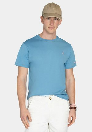 Joven con gorra beige, camiseta azul clara, pantalones blancos y reloj de pulsera con una pulsera rosa, de pie frente a un fondo liso.