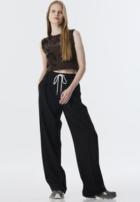 Top corto de punto marrón con patrón texturizado, combinado con pantalones negros holgados que tienen cintura con cordón. Modelo lleva zapatillas grises.