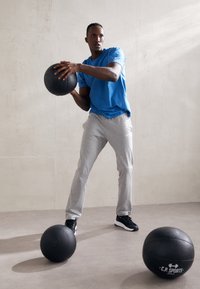 Melna medicīnas bumba rokās, tērpies zilā sporta krekla un pelēkās sporta biksēs, stāv uz gludas grīdas, blakus vēl divām melnām bumbām.