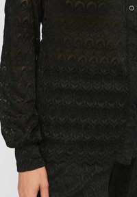 Must mustriga blus, millel on tekstuuriga lehe muster, pikad varrukad, nööbikinnitus ja laineline äär, mis lisab detailirohkusele.